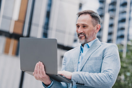 Confident mature businessman in a modern cityscape uses a sleek laptop outdoors smiling while working with clients in a stylish professional settingの写真素材