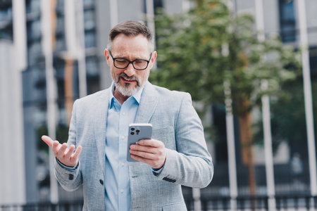 A mature businessman in a light blue suit uses a smartphone while standing in a modern city streetの写真素材