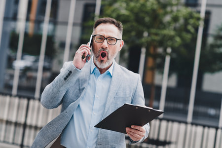 Businessman on the move in a modern city speaks on the phone while holding a clipboard outdoors in a professional settingの写真素材