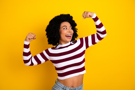 Young woman in striped casual outfit flexing arms confidently on yellow background, showcasing happiness, beauty, and strengthの写真素材