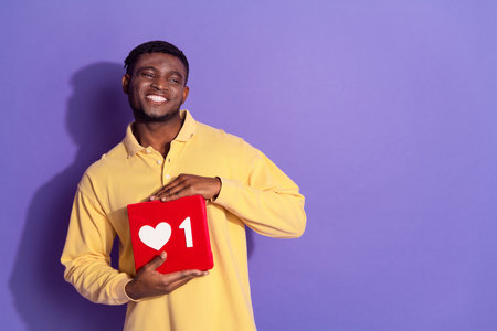 Photo of pretty young male hold social media like icon look empty space wear trendy yellow outfit isolated on purple color backgroundの写真素材