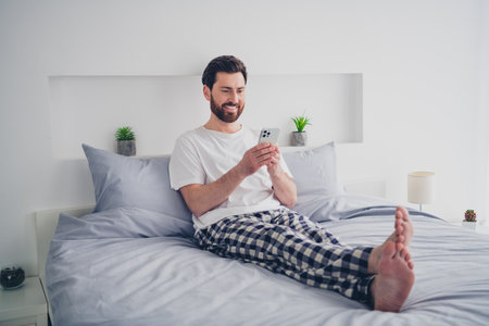 Photo of attractive funny young man nightwear pajama sitting bed chatting modern device indoors house apartment roomの写真素材