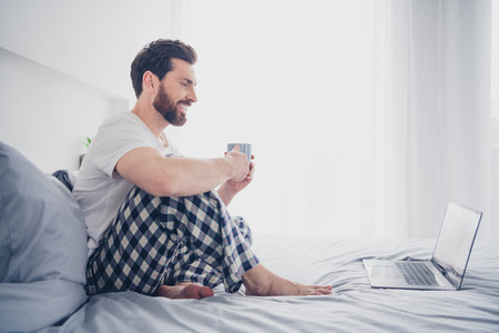 Photo of attractive funny young man nightwear pajama sitting bed drinking coffee chatting modern device indoors house apartment roomの写真素材