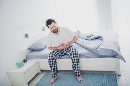 Photo of unhappy stressed young man nightwear pajama sitting bed feeling stomach ache indoors house apartment roomの写真素材