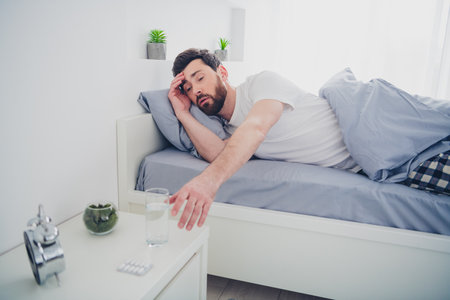 Photo of funny tired young man dressed nightwear lying bed want drink water indoors house apartment roomの写真素材