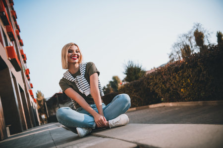 Full length photo of attractive young cheerful lady sit concrete border wear khaki garment tied shoulders sweater have fun outside outdoorsの写真素材
