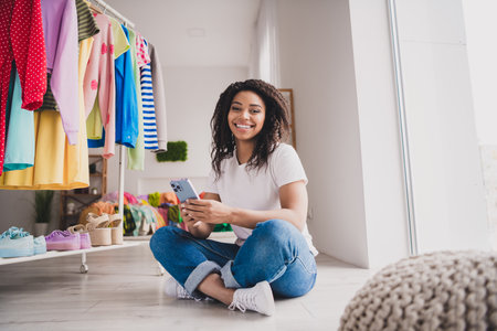 Young woman enjoys online shopping on smartphone in cozy living room with clothes rack on sunny dayの写真素材