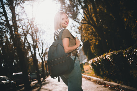 Photo of pretty young blonde woman backpack turn around wear khaki clothes tied shoulders sweater walk outside urban city parkの写真素材