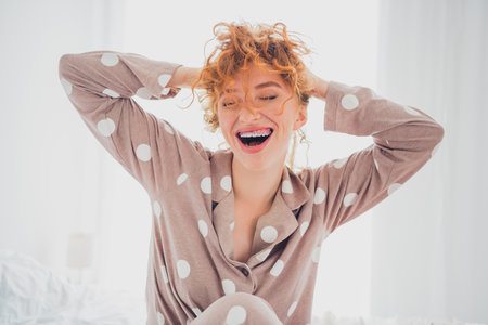 Charming young woman with red hair smiling in polka dot sleepwear during a joyful morning in a bright bedroomの写真素材