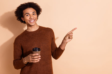 Young African American man in a casual sweater pointing with a cheerful smile against a beige background, holding a coffee cupの写真素材
