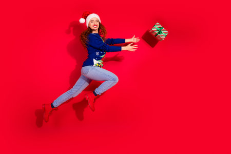 Full length photo of charming positive girl wear print sweater jumping high celebrating catching xmas gifts empty space isolated red color backgroundの写真素材