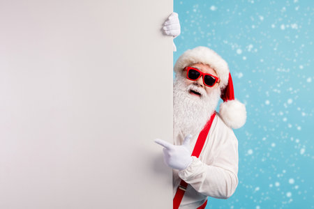 Photo of pensioner grandfather grey beard direct fingers empty space promoting seasonal discounts wear santa costume suspenders spectacles white headwear isolated blue color backgroundの写真素材