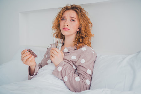 Young woman with red hair sitting in bed holding glass and pills, wearing cozy sleepwear in bright bedroomの写真素材