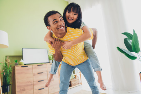 Photo of lovely sweet multinational couple wear t-shirts have fun time together hold hands piggyback indoors flatの写真素材