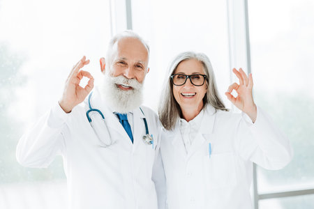 Two friendly elderly doctors in white coats smile at the camera in a bright clinic giving an ok gesture reflecting teamwork care and trust in health careの写真素材