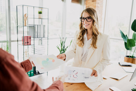 Professional businesswoman in elegant suit handing over documents in a modern office setting during daytimeの写真素材