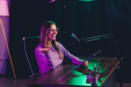 Woman in casual attire conducting a podcast with a microphone in a neon-lit home office at nightの写真素材