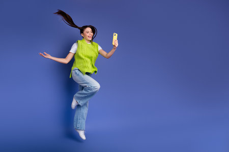 Young woman in neon green vest jumps against blue background smiling while taking a selfie with a...