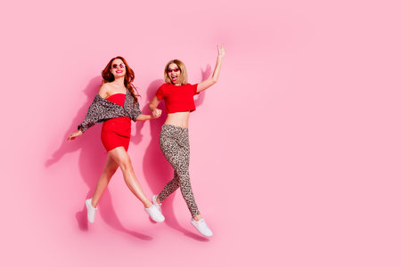 Full length photo of two lovely young ladies jump running hold hands dressed stylish red leopard print garment isolated on pink backgroundの写真素材