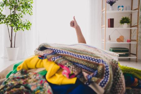 Joyful african american girl surrounded by colorful clothes showing thumbs up at home