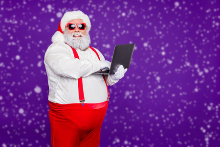 Profile side view portrait of his he nice confident cheerful cheery bearded Santa holding in hands laptop typing e-mail letter isolated on bright vivid shine vibrant violet lilac backgroundの写真素材