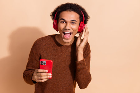 Young african american male enjoying music with headphones, holding smartphone, standing against beige backdropの写真素材