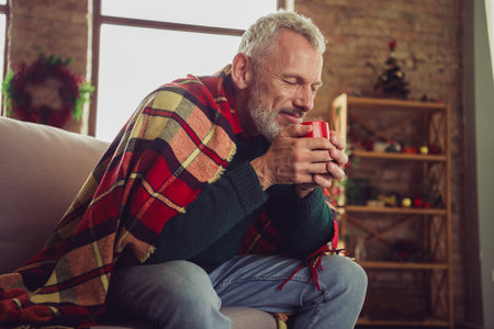 Portrait of nice aged man sit sofa covered blanket smell coffee wear sweater new year xmas time flat indoorsの写真素材