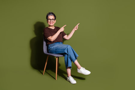 Full size photo of lovely pensioner lady sit chair point empty space dressed stylish brown garment isolated on khaki color backgroundの写真素材