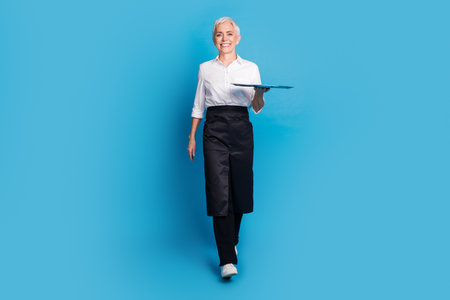 Full body photo of attractive senior woman walk carry tray small business cafe owner dressed waitress uniform isolated on blue backgroundの写真素材