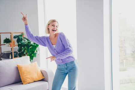 Young woman in casual attire dances joyfully near a large window inside her cozy living room on a bright sunny dayの写真素材