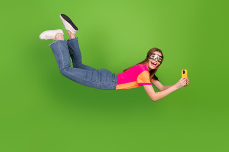 Young woman in bright casual outfit wearing goggles flies in mid air while taking a selfie with a...