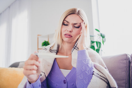 Young woman feeling unwell while checking her temperature at home, wrapped in a cozy knitted...
