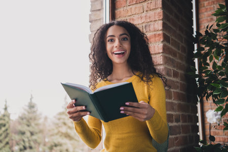Young woman reading enthusiastically at home near a brick wall, capturing the joy of learning in natural light and a cozy setting.の写真素材