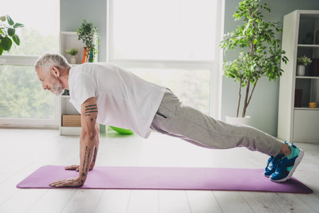Elderly man practicing fitness exercises on a yoga mat at home in bright daylight with focus on...