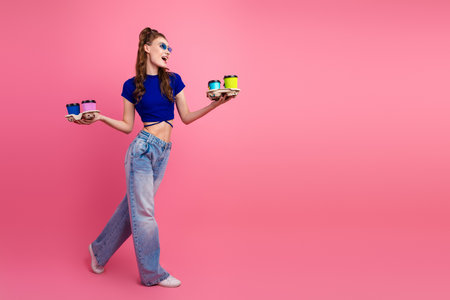 Happy young woman in casual outfit holding colorful coffee cups standing against a vibrant pink...