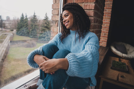 Charming young woman enjoying a relaxing weekend at home in a cozy blue sweater by the windowの写真素材