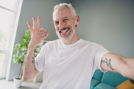 Energetic mature man in a white t-shirt smiling indoors and making an okay gesture with his tattooed...