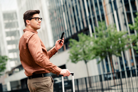 Confident young businessman uses his smartphone while traveling in a vibrant urban cityscape for professional pursuitsの写真素材