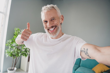 Happy mature man in white t-shirt giving thumbs up while smiling warmly in a bright home living room...