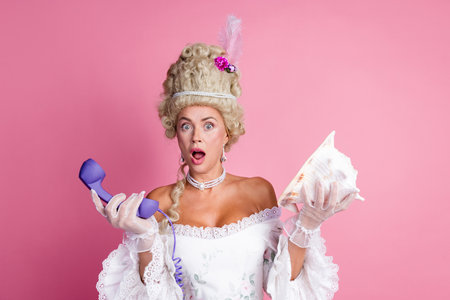 Elegant woman in vintage baroque attire expresses surprise while holding a phone and a shell against a vibrant pink backgroundの写真素材