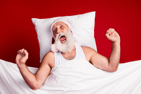 Festive sleepy Santa man yawns in bed with pillow and blanket in a red themed Christmas moodの写真素材