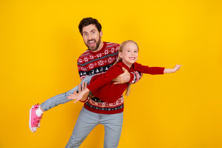 Cheerful father holding cheerful daughter in a playful airplane pose, with festive sweaters on a bright yellow background.の写真素材