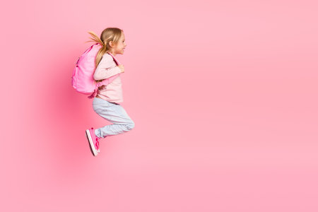 Lovely young girl with pink backpack jumping joyfully in a stylish outfit on a matching pink background showcasing happy energyの写真素材