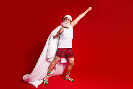 Festive Santa figure strikes a bold pose in a white tank top red plaid shorts and cape against a striking red backdropの写真素材