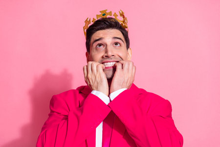 Young man in pink suit wearing gold crown looking amused against vibrant pink background expressing quirky emotionsの写真素材