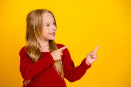 Funny charming young girl in red knit sweater points at herself against bright yellow background in...