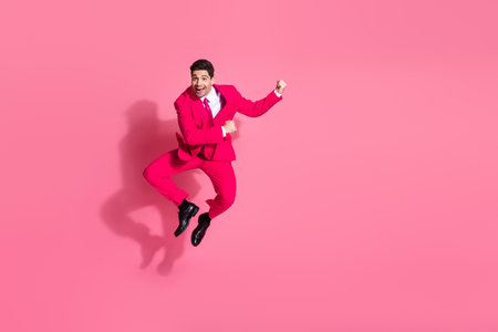 Energetic young man in a pink suit jumping joyfully on a solid pink background, inspiring fun and...