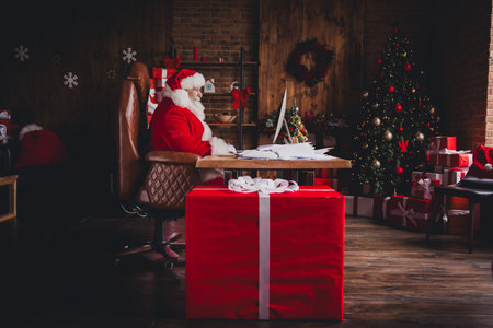 Santa at his desk in a cozy Christmas workshop surrounded by gifts and a glowing tree full of lights and holiday magicの写真素材