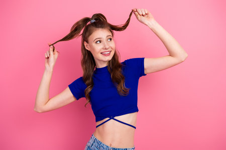 Charming young brunette woman posing with playful hairstyle against pink background, wearing casual blue top and jeansの写真素材