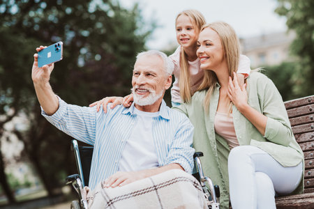 Heartwarming family moment as grandpa in wheelchair smiles with daughter and granddaughter on a sunny park bench together outdoorsの写真素材